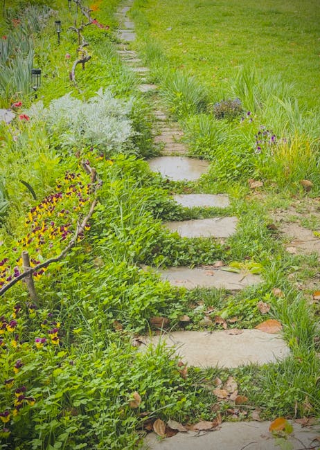 A narrow garden pathway constructed from irregularly shaped stepping stones set into the soil, bordered by lush, green plants and vibrant flowering shrubs. On the left side, there is a decorative wood